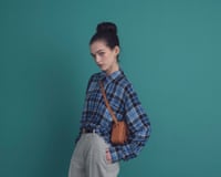 Female model wearing blue plaid shirt half-tucked into grey trousers with belt and small crossbody bag