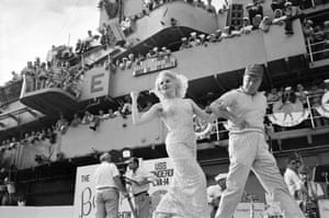Actor Carroll Baker snaps her fingers at sailors cheering from the bridge as Bob Hope leads her across the stage on the flight deck of the USS Ticonderoga, December 1965
