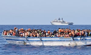 Migrants in a wooden-hulled ship wearing lifejackets provided by HMS Bulwark, seen in the background