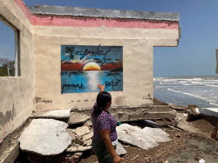 A woman stands in front of the old school building, with walls missing and the tide lapping next to it