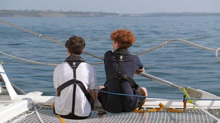 Two teenage boys, pictured from behind sitting on the netting of a boat, look out across the sea towards the shore