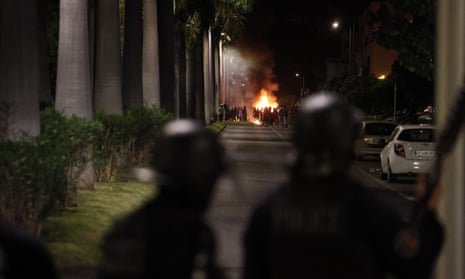 Riot police stand guard and watch a group of people standing around a fire.