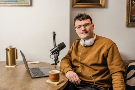 Nick Hilton, wearing a brown sweatshirt, with white Bose headphones around his neck, sitting at a desk with a laptop, a large microphone, a coffee pot and a mug on a coaster