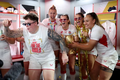 Female rugby players in white T-shirts and shorts celebrate with their trophy, cheering and laughing; one has her arm raised