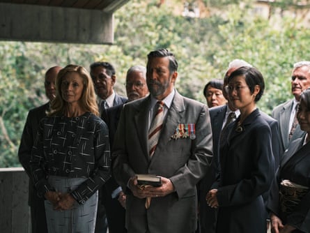Heather Mitchell and Ciaran Hinds stand at the front of a small group of people on a balcony with trees in the background. He is wearing a grey suit, with war medals pinned to its left breast.