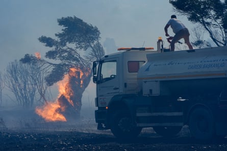 A man sprays water from a tanker to extinguish burning olive trees amid dark smoke in İzmir, Turkey.