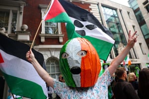 A person in a papier maché balaclava in the colours of the Irish flag waves a Palestinian flag