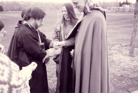 Man in robe ministers to man in cape, people smiling on lawn.