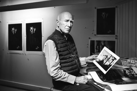 An elderly white man with a sombre face stands in a studio holding a black-and-white photograph with other prints on the wall behind him