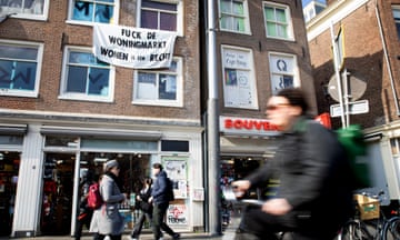 A banner hanging from the upper windows of  building on a busy street in Amsterdam bears the message ‘fuck the housing market’. Pedestrians and cyclists pass by in the foreground