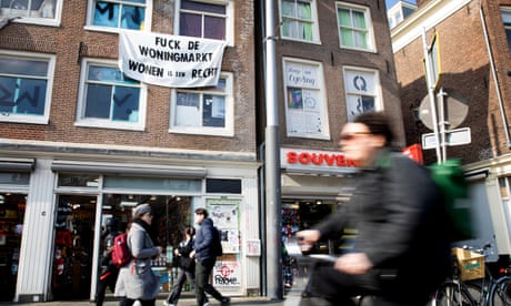 A banner hanging from the upper windows of building on a busy street in Amsterdam bears the message ‘fuck the housing market’. Pedestrians and cyclists pass by in the foreground