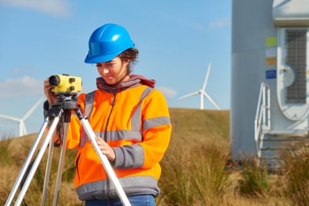 A female windfarm engineer in orange gi-vis jacket and blue hard hat looks through a builder's level as she prepares construction of a wind turbine. In the background more turbines can be seen.