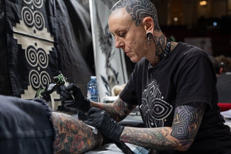 Woman with a shaved, tattooed head and arms tattoos someone’s leg