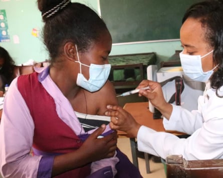 A woman holding a syringe inserts the needle into a schoolgirl’s arm in a classroom