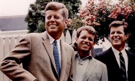 A color-tinted photo of three smiling white men wearing casual suits and standing in a row, looking in the same direction, in front of a pink rosebush and a white picket fence.