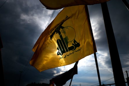 A yellow Hezbollah flag, bearing green writing in Arabic and a gun symbol, is held up against a dark, cloudy sky.