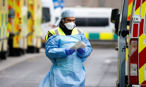 A medical worker wearing a face mask and protective clothing walks beside ambulances outside the emergency department of the Royal London Hospital