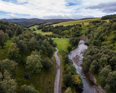 An aerial view of cyclists on a road, with trees, green fields and mountains in the distance