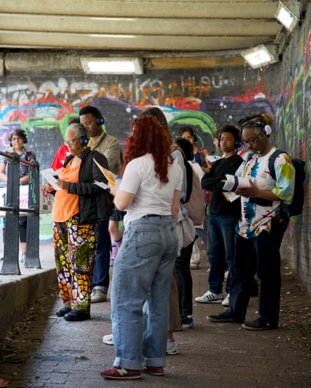 A group of people wearing large over-ear headphones stand under a concrete bridge or tunnel covered in colourful graffiti