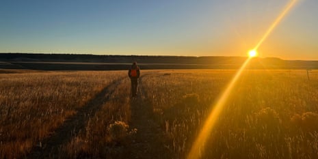 A person wearing an orange vest walks alone along a dirt path through a golden grassy field at sunset. The sun is low on the horizon, casting a bright beam of light across the landscape under a clear sky.
