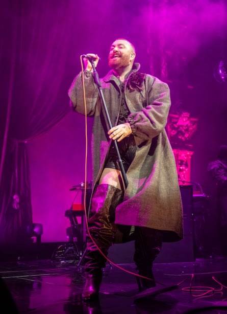 Smith holding a stand mic on stage, wearing a long brown coat and long boots