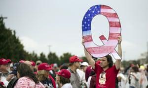 A protester holding a Q sign decorated with the American flag waits in line with others to enter a Trump rally