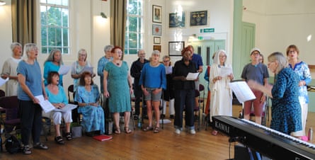 A group of 21 men and women stand in an arc in a village hall. A choirmaster is conducting proceedings next to a piano