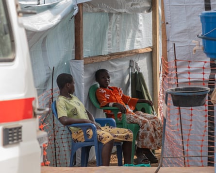 Two girls seated together under a fabric shelter by a van