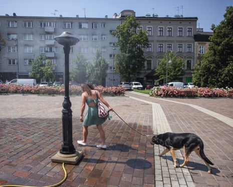 A woman in a green dress walks a dog under a water sprinkler in a public square