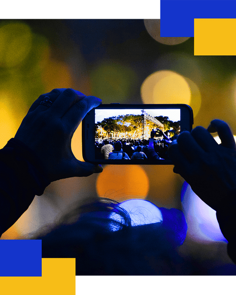 Silhouetted hands hold up a phone recording a concert crowd at night, surrounded by blurred lights