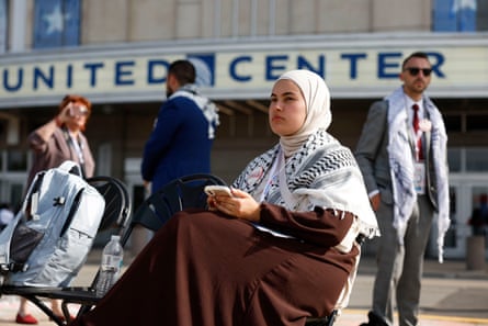 Woman wearing keffiyeh as head scarf and long brown dress sits in folding chair.
