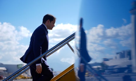 Rubio walks up steps, reflected in body of plane with white clouds in blue sky