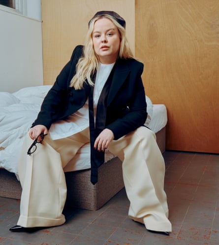 Actor Nicola Coughlan sitting on a bed in black jacket and hat, white top and cream trousers