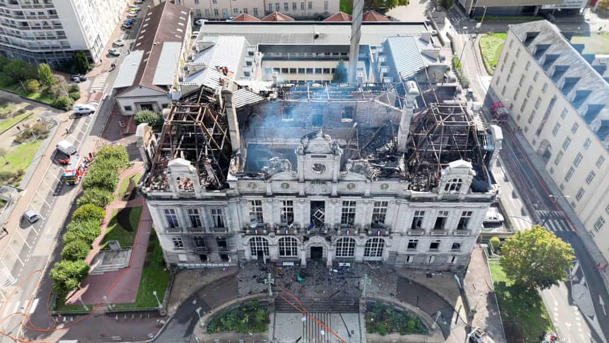 Overhead view showing the burned-out roof of the city hall with smoke still rising. The front facade and side walls remain.