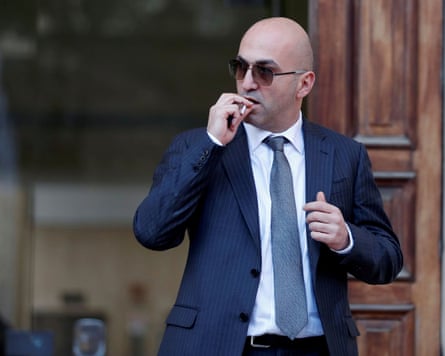 Yorgen Fenech puts a cigarette to his mouth as in front of a large wooden door