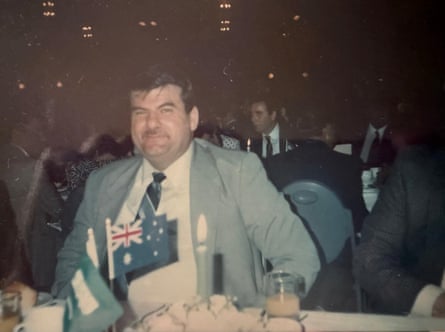 Abdul Ghazal, an older Lebanese Australian man in a suit seated at a dining table with many suited people in the background seated behind him. An Australian flag is visible in the foreground.