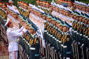 Soldiers in ceremonial uniforms, some green and some white, stand to attention while another solider adjusts one of their peaked caps