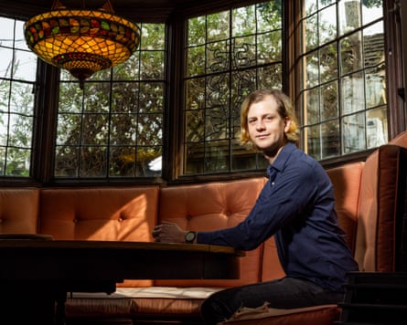 Man with long hair wearing a blue shirt, sat at a banquette by a window in a nice house