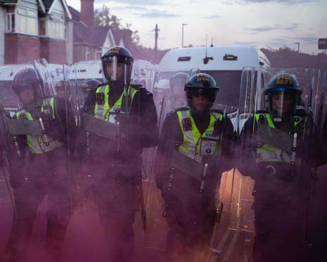 Police officers in riot gear walk form a line amid smoke from flares