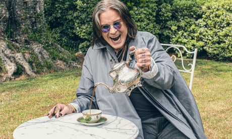 Ozzy Osbourne in a garden, sitting at a white iron table pouring tea from an ornate silver teapot, with his trademark cloak, black nail varnish, round blue sunglasses and open-mouthed grin