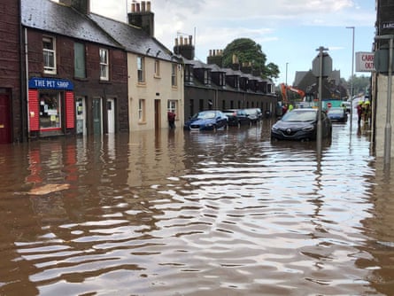 A flooded Scottish street, with cars standing in water, their wheels just breaking the surface. A row of houses in the background with one shop called The Pet Shop.