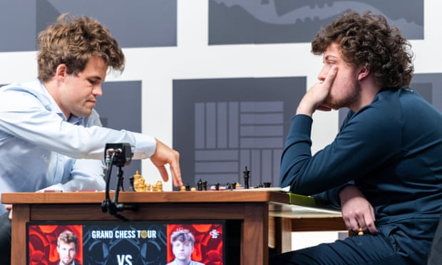 Two men sit at a chess board. The one on the left is in the middle of a move, the one on the right is studying him carefully.