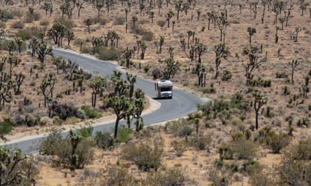 A two-lane asphalt road curves through a barren landscape dotted with scrubby, unusual-looking trees, with a beige camper van driving through it.