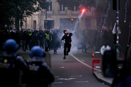 A protester wearing a face mask and black hoodie throws a smoke flare towards gendarmes in a street in central Bordeaux. The police face the protesters and wear blue helmets.