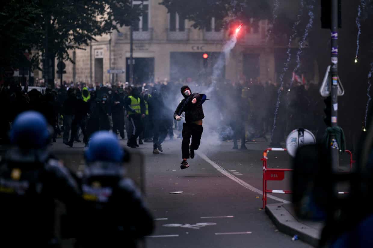 A protester wearing a face mask and black hoodie throws a smoke flare towards gendarmes in a street in central Bordeaux. The police face the protesters and wear blue helmets.