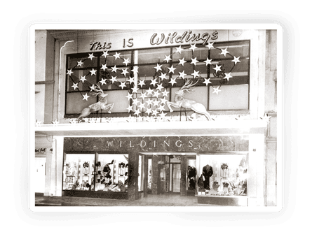 An archival black-and-white photograph showing Wildings department store from the street. The Christmas display on the first floor level shows two reindeer with many gold stars above their antlers