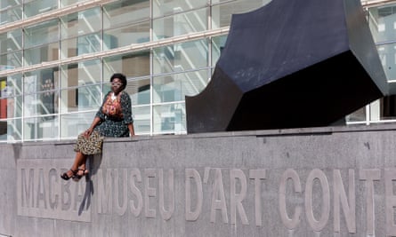 Elvira Dyangani Ose sitting on a wall bearing the museum's logo and name in relief along its length, with a dark, jagged abstract culture to her left on top of the wall