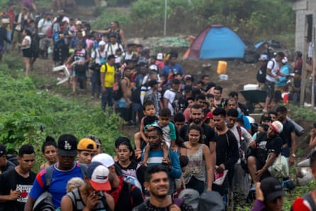 A large crowd of people carrying children and belongings passes by a small camping tent in the jungle