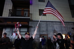 A man in a baseball cap holding up both arms, with a US flag in one hand