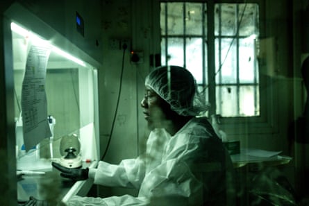 A medical worker in a hairnet, black latex gloves and a white medical gown sits in front of a protective screen